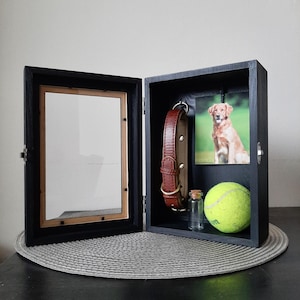 Peut inclure: Un coffret souvenir pour animaux de compagnie en bois noir avec une porte à charnière. À l'intérieur, un collier de chien en cuir marron, une photo d'un golden retriever, une balle de tennis et une petite bouteille en verre sont exposés. L'autre côté a un cadre photo.