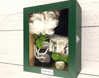 May include: A green wooden shadow box with a glass front. Inside the box are white flowers, a black fabric bag, a pearl necklace, a black number "30", a photo of a couple, and a small white flower. The box has a silver latch and a label that reads "30th Anniversary".