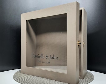 May include: A light gray wooden box with a glass front and a hinged lid. The box has a gold latch and is engraved with the text "Danielle & John Oct 20, 2018".