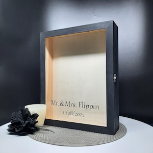 May include: Black wooden shadow box with a glass front. The box has a hinged door and is personalized with the text "Mr. & Mrs. Flippin 11/08/2022".