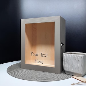 Peut inclure: Une boîte à ombre en bois gris avec une face en verre. La boîte a un couvercle à charnières et une serrure. L'intérieur de la boîte est doublé de bois. La boîte est posée sur un set de table gris. Le texte "Your Text Here" est imprimé à l'intérieur de la boîte.