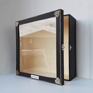 May include: Black wooden shadow box with a glass front and a hinged door. The box has a distressed finish and decorative metal accents. The inside of the box is lined with a light brown fabric. The box has a label that says "Your Text Here".