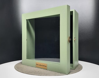 May include: A sage green shadow box with a glass front and a brass latch. The box is open and the inside is black. The box has a brass plaque on the bottom with the text "Your custom text here".