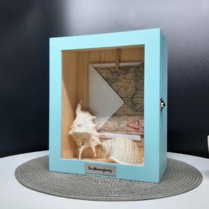 May include: A light blue wooden shadow box with a glass front. Inside the box are two seashells, a white envelope, and a vintage map. The box has a silver plaque on the bottom that reads "The Moominfamily".