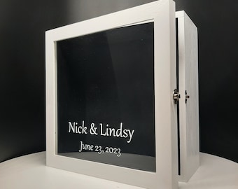 May include: White wooden box with a glass front and a black background. The box has a hinged door and a silver latch. The glass front has the text "Nick & Lindsey June 23, 2023" in white lettering.