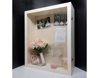 May include: A white shadow box with a glass front, containing a bouquet of pink and white flowers, two champagne flutes, a small pink box, a candle, and a framed wedding vow card. The shadow box also includes four photos of a bride and groom.