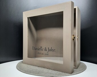 May include: A light gray wooden shadow box with a glass front. The box has a hinged door and a brass latch. The box is engraved with the text "Danielle & John Oct 20, 2018".