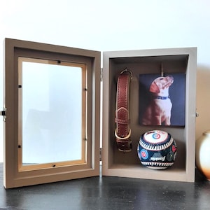 May include: A light brown wooden shadow box display case with a hinged glass door. Inside, a brown leather pet collar hangs next to a square photograph of a dog and a white baseball with red and blue stitching and various team logos printed on it.