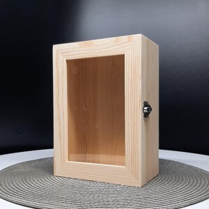 Peut inclure: Une vitrine en bois avec une façade en verre et un loquet en métal. La vitrine est en bois naturel et a une couleur brun clair.