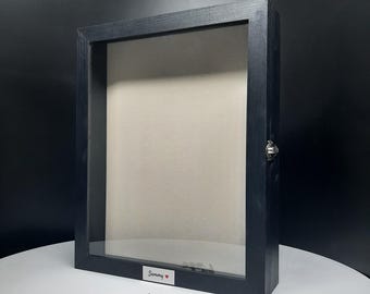 May include: A black wooden display case with a glass front and a light-colored interior. The case has a hinged door with a metal clasp and the name "Sammy" is printed on the bottom. Several small silver-colored objects are in front of the case.