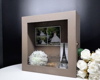 May include: A light brown wooden shadow box with a glass front. The box contains a wedding photo, a small Eiffel Tower figurine, a white flower, and a small box. The box is personalized with the text "Williams family est. 2012".