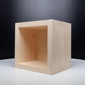 May include: A square, open-front wooden box crafted from light-colored, unfinished pine wood. The structure features visible finger joints at the corners, showcasing the natural grain patterns and texture of the wood in a simple, boxy design.