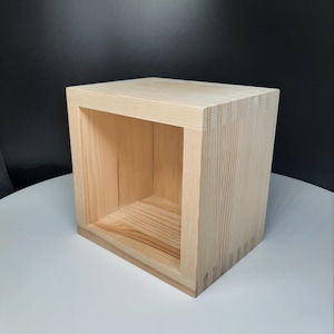 May include: A light-colored, wooden cube-shaped shelf with an open front. The shelf is constructed with visible dovetail joints at the corners. The wood grain is visible, and the shelf is set against a white and black background.
