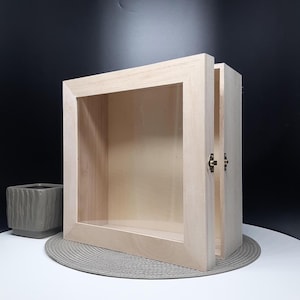 May include: A light-colored wooden display box with a glass front and a hinged door. The door is partially open, revealing the interior. The box is rectangular and sits on a gray placemat. The hardware is a brass color.