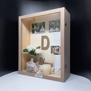 May include: A wooden shadow box with a white background. The box contains a bouquet of white roses, a wooden sign that says "Home", a gold letter "D", and several photographs. The box is decorated with a ribbon and a candle.