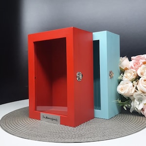 May include: Two wooden shadow boxes with glass fronts. The box on the left is red and has a silver plaque with the text "The Moominfamily". The box on the right is light blue.