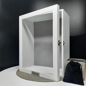 May include: White wooden shadow box with a glass front and a gray backing. The box has a silver plaque with the text "The McManus Family" on the bottom.