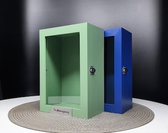 May include: Two wooden shadow boxes with glass fronts. The box on the left is green and the box on the right is blue. Both boxes have a silver latch and a silver nameplate. The nameplate on the green box reads "The Moominfamily".