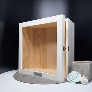 May include: White wooden shadow box with a hinged door. The box has a clear glass front and a wooden back. The box is sitting on a gray surface. The box has a small metal plate on the bottom with the text "The Memory Jar" engraved on it.