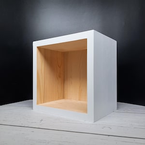 May include: A white, square wooden shelf unit with a natural wood interior. The cube-shaped shelf is painted white on the exterior and sits on a light-colored wooden surface against a black background. The shelf is designed to be a storage or display unit.