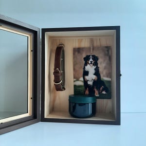 May include: A wooden pet memorial box with a hinged door. Inside, a brown leather dog collar hangs next to a framed photo of a Bernese Mountain Dog. A small, dark blue jar sits below the photo.