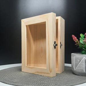 May include: A small, open wooden display cabinet with a glass front. The cabinet is made of light-colored wood and has a hinged door with a metal latch. The cabinet is empty and sits on a gray placemat. A potted plant is in the background.
