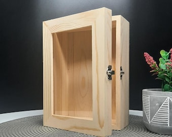 Peut inclure: Un petit présentoir en bois ouvert avec une façade en verre. L'armoire est en bois de couleur claire et possède une porte à charnière avec un loquet en métal. L'armoire est vide et repose sur un dessous de plat gris. Une plante en pot est en arrière-plan.