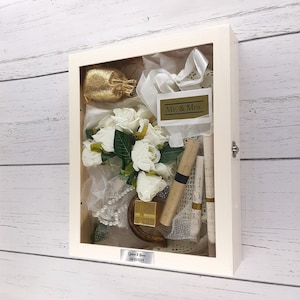 May include: A white wooden shadow box with a glass front, containing a white bouquet of roses, a gold bag, a pearl necklace, a gold gift box, and a scroll. The box has a label that says "Mr. & Mrs." and a personalised label with the date "08/05/2024".