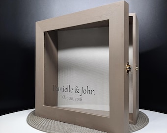May include: A light brown wooden shadow box with a glass front. The box has a hinged door on the right side. The glass front has the text "Danielle & John Oct 20, 2018" printed on it.