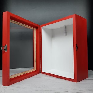 May include: A red wooden display case with a glass front and a hinged door. The case is open and the interior is white.
