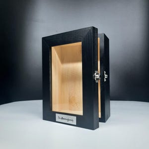 Puede incluir: Una vitrina de madera negra con frente de cristal y puerta abatible. El interior es de madera clara. Una pequeña placa metálica en la parte delantera dice "The Moominfamily". La vitrina está abierta.
