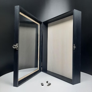 May include: Black wooden shadow box with a glass front and a white backing. The box is open and the glass front is facing the camera. The box is sitting on a white surface.