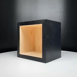 May include: A square, open-front wooden shelf in a dark charcoal finish. The interior of the shelf is natural wood, creating a contrast. The shelf is designed to be a decorative storage unit.