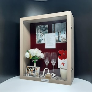 May include: A shadow box display with a light wood frame. Inside, a bouquet of white roses, two champagne flutes, and wedding photos are arranged against a burgundy backdrop. Decorative elements include a "Love" sign and a small gift box.