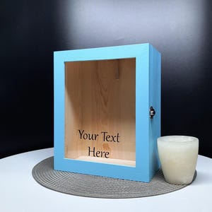 May include: A light blue wooden box with a glass front. The box has a small metal latch on the side. The glass front has a white background with the text "Your Text Here" printed in black.