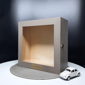 May include: A wooden display box with a clear front panel and a small metal clasp. The box is painted in a neutral gray color and has a small gold-colored plate that reads "Your custom text here". A white toy car sits in front of the box.