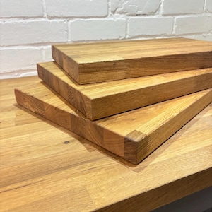 May include: Three rectangular wooden chopping boards stacked. The boards are made of light brown wood with visible grain and are placed on a matching wooden surface. The boards have a natural, unfinished appearance, suitable for kitchen use.