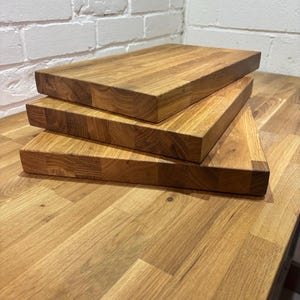 May include: Three wooden chopping boards stacked. Each board is rectangular with rounded edges and a natural wood grain finish. The boards are displayed on a wooden surface, showing the natural wood grain.