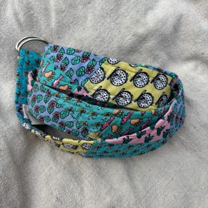 May include: A fabric belt featuring a colorful patchwork design with patterns of fishing reels, flies, and leaves in shades of yellow, teal, pink, and blue. The belt is secured with two silver-toned metal D-rings at one end, providing a functional closure.