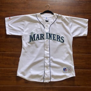 May include: White baseball jersey with a navy blue trim and the word "Mariners" in green and gold lettering with a star above the word.