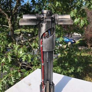 Puede incluir: Una réplica de sable de luz detallada y desmontada con un acabado plateado y negro. El sable de luz está hecho de metal y tiene un cable rojo que lo atraviesa. El sable de luz está sobre una superficie blanca.