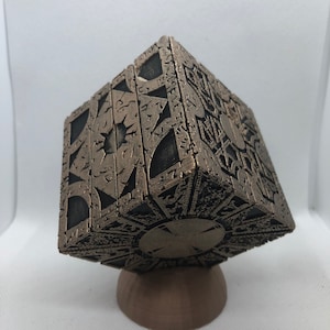 May include: A detailed, intricate, bronze-coloured metal cube puzzle with a circular design on the bottom. The cube is sitting on a small, round, bronze-coloured base.