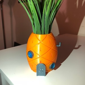 May include: A 3D printed pineapple-shaped planter with a grey door and blue windows. The planter is filled with green artificial grass.