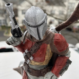 May include: A 3D printed action figure of a Mandalorian warrior. The figure is wearing a silver helmet with a T-shaped visor, a red and brown chest plate, and a brown belt. The figure is holding a blaster pistol in its right hand.