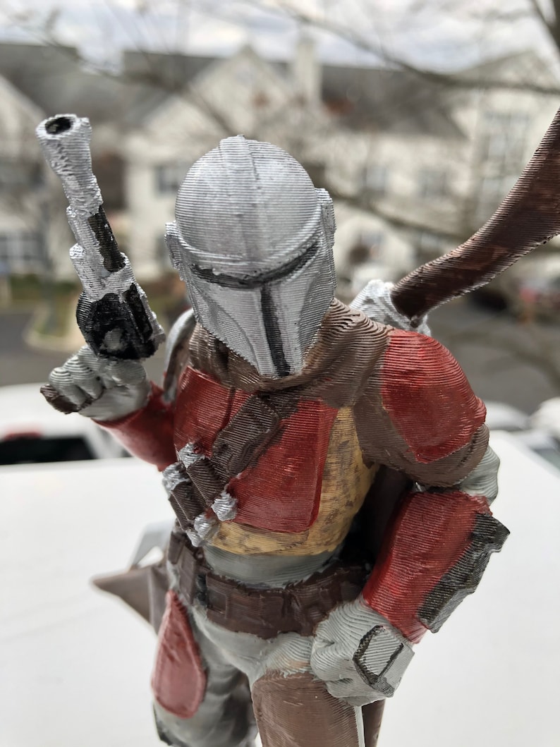 May include: A 3D printed action figure of a Mandalorian warrior. The figure is wearing a silver helmet with a T-shaped visor, a red and brown chest plate, and a brown belt. The figure is holding a blaster pistol in its right hand.
