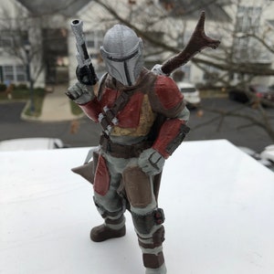May include: A 3D printed action figure of a Mandalorian warrior. The figure is wearing a gray helmet with a T-shaped visor, a brown and red armor, and a brown cape. The figure is holding a blaster pistol in one hand and a staff in the other.