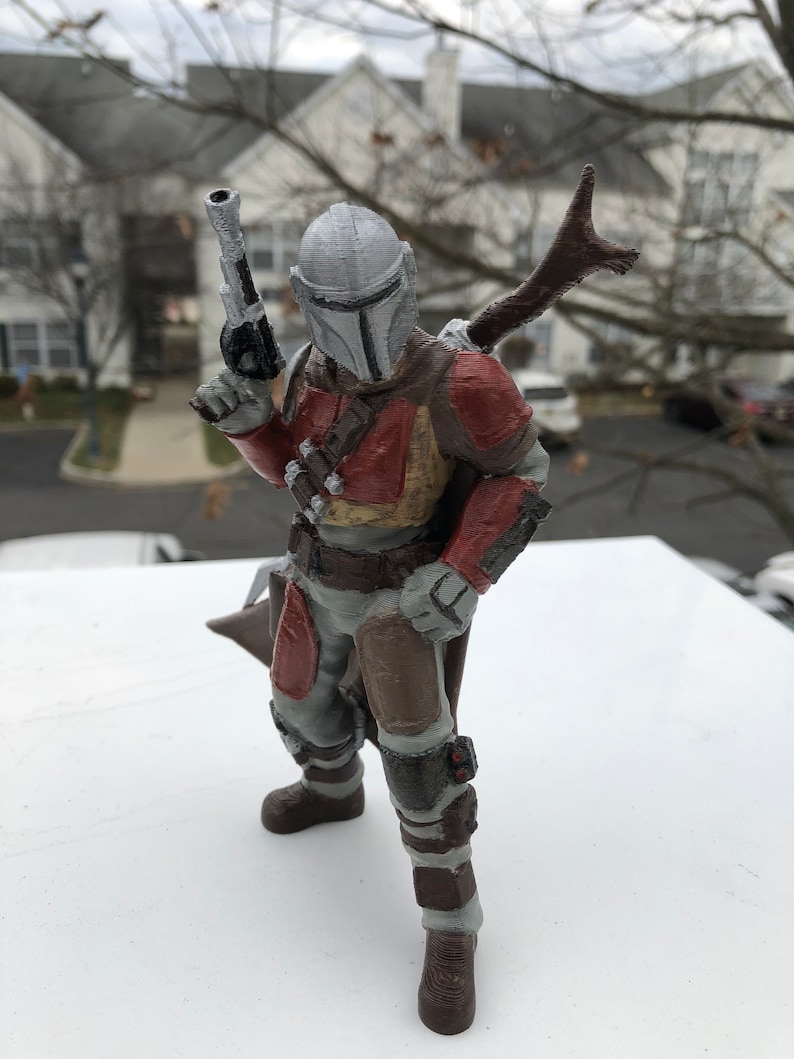 May include: A 3D printed action figure of a Mandalorian warrior. The figure is wearing a gray helmet with a T-shaped visor, a brown and red armor, and a brown cape. The figure is holding a blaster pistol in one hand and a staff in the other.