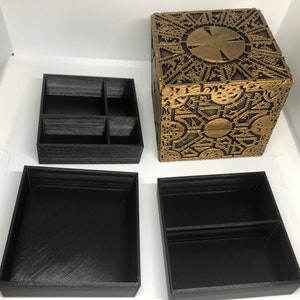 Może przedstawiać: Czarne pudełko wydrukowane w 3D ze skomplikowanym wzorem pomalowanym na złoto. Pudełko jest otwarte i ma trzy mniejsze przegródki w środku. Pudełko jest repliką kultowej pudełka z łamigłówką z filmu "Siedem".