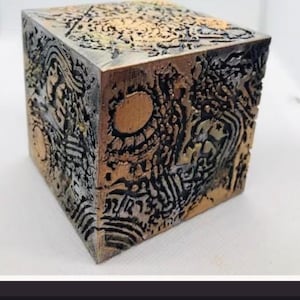 Op de afbeelding: Een metalen kubus met een complex, verhoogd reliëfpatroon met een donkere, geoxideerde afwerking en gepolijste bronskleurige accenten. Het oppervlak toont ingewikkelde geometrische vormen, cirkelvormige motieven en kronkelende lijnen op de zijden.