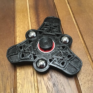 Puede incluir: Spinner de plástico negro con un diseño de Darth Vader y cuatro rodamientos plateados. El spinner tiene un centro rojo y blanco.